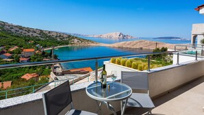 Un balcon avec une table en verre, deux chaises et une bouteille de vin. La vue comprend une étendue d’eau, une côte rocheuse et des montagnes lointaines.