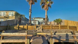 A wooden boardwalk leading to a beach area with palm trees and a building in the background.