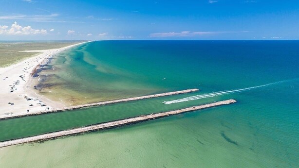 An aerial view of a beach with clear waters, a long pier, and a sandy shoreline.
