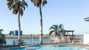 A pool area with two palm trees, a pool, and a view of the sea.