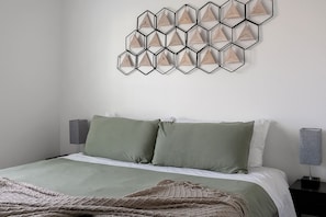 Un dormitorio ordenado con una cama, dos almohadas, una colcha y un accesorio de luz geométrico montado en la pared.