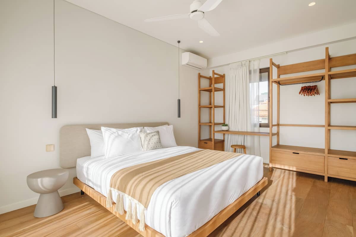Un dormitorio moderno con una cama grande, ventilador de techo, equipo de aire acondicionado, armario de madera y mesita de noche.