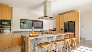 A modern kitchen with wooden cabinets, a central island with stools, stainless steel appliances, and a large window.