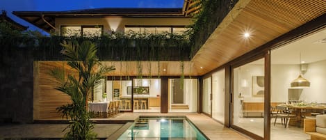 A modern outdoor pool area with a wooden deck, a dining area with a table and chairs, and a kitchen visible through glass doors.