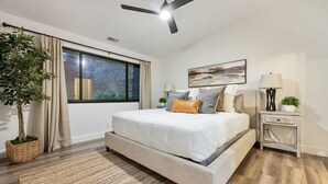 A modern bedroom with a large bed, a ceiling fan, a window with curtains, a bedside table with a lamp, and a potted plant.