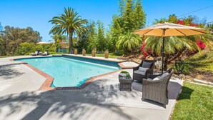 A swimming pool with lounge chairs and an umbrella, surrounded by greenery and palm trees.
