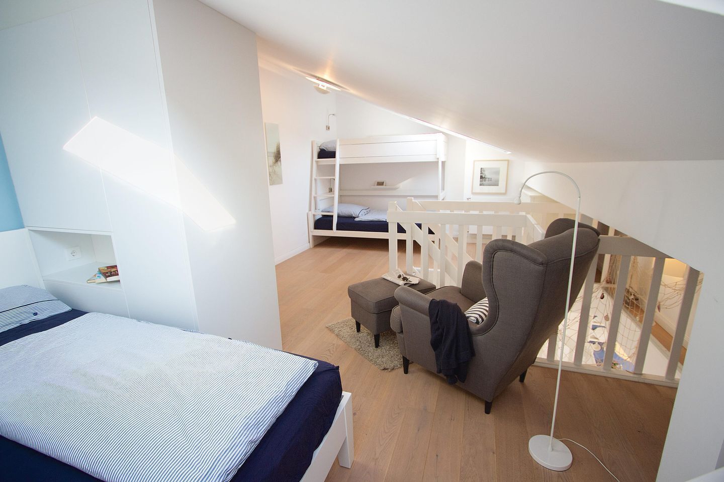 Een moderne slaapkamer met een stapelbed, een groot tweepersoonsbed, een grijze fauteuil en een vloerlamp.
