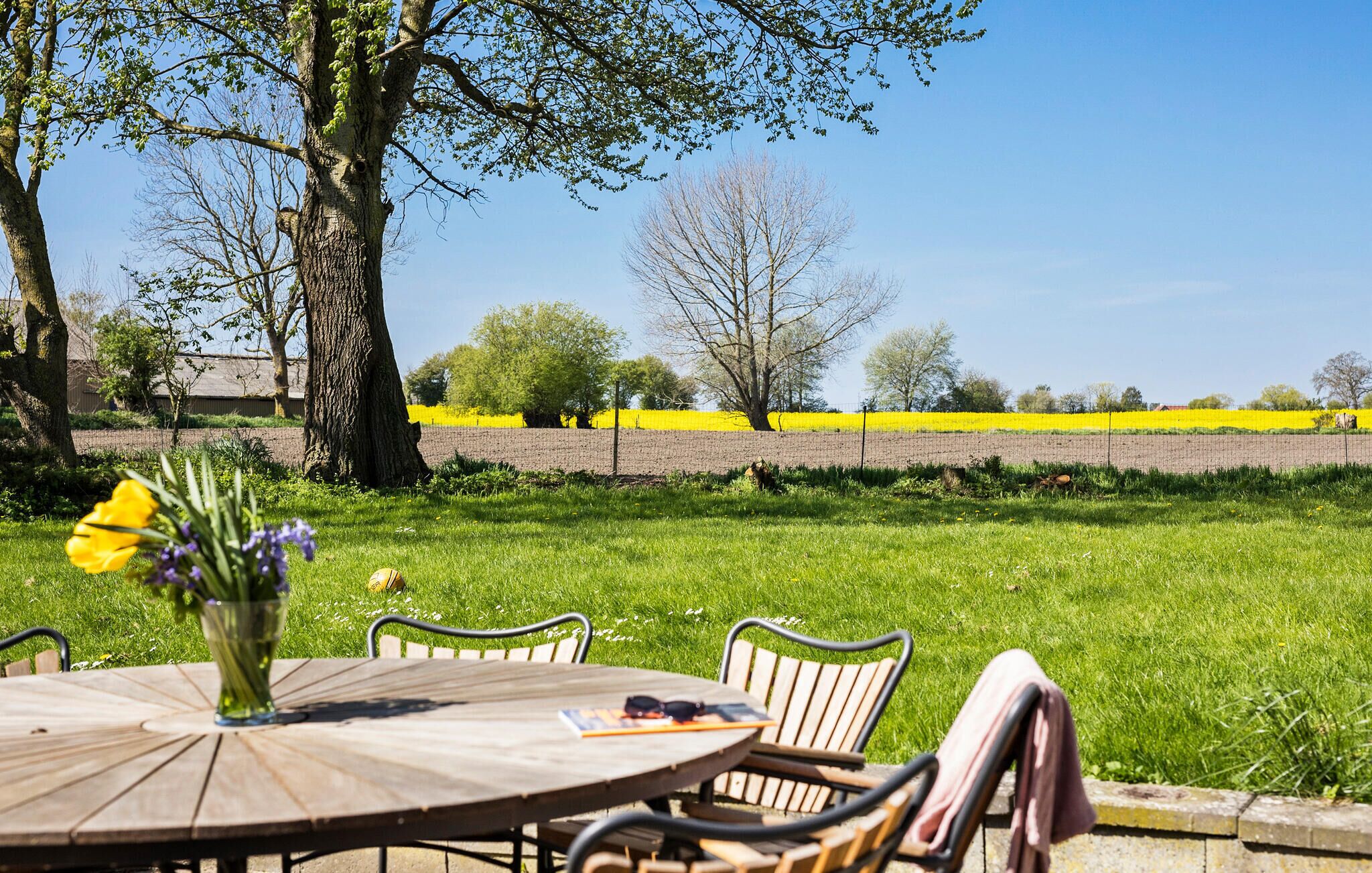 An outdoor seating area with a round wooden table and metal chairs, surrounded by a grassy field and trees.
