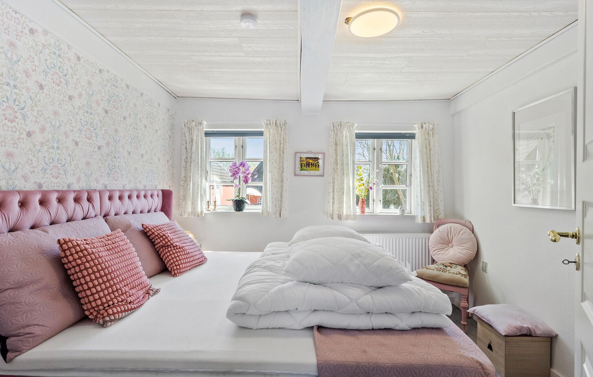 Une chambre avec un grand lit, une tête de lit rembourrée de couleur rose et une fenêtre avec des rideaux.