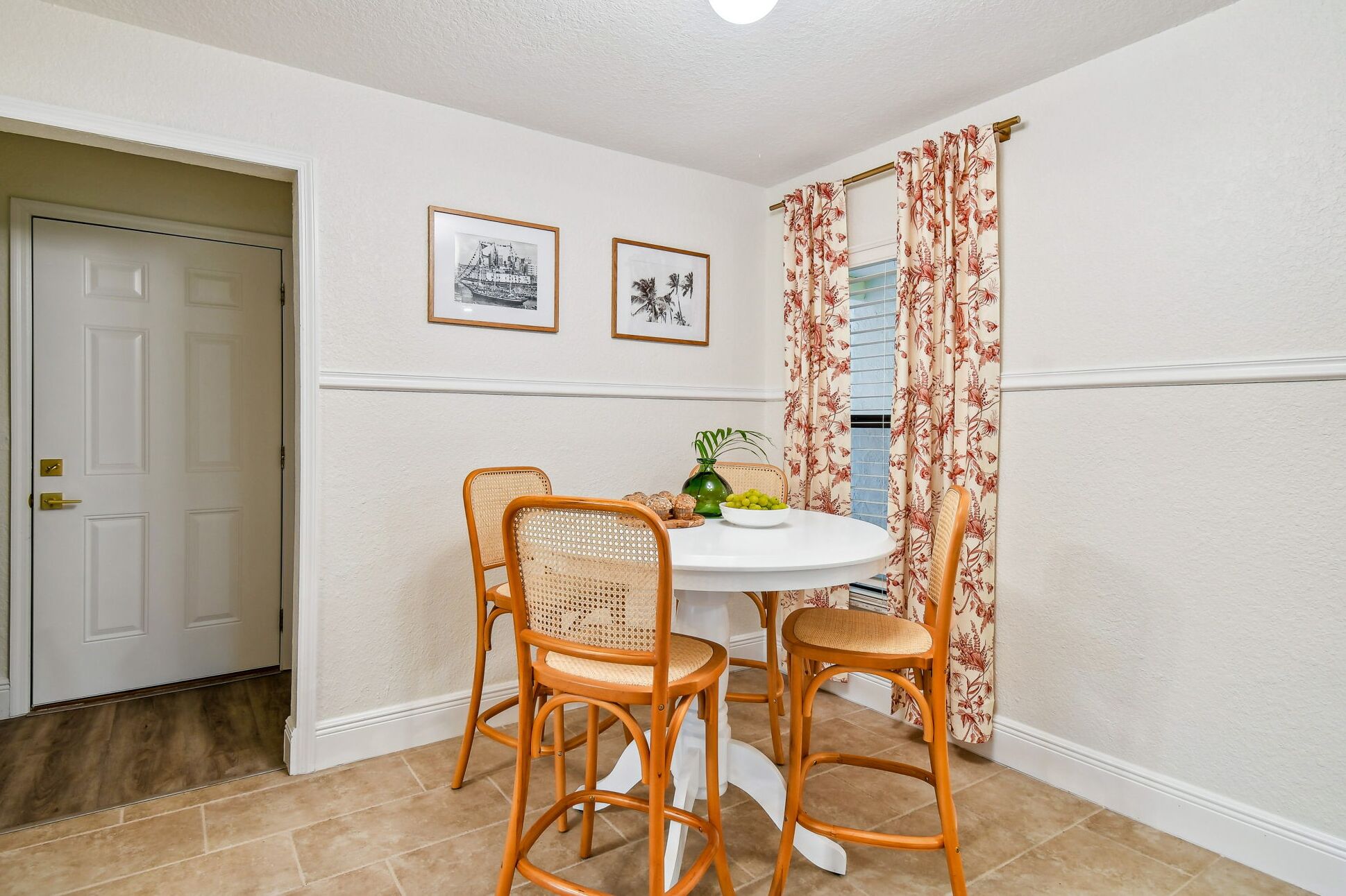 Een eethoek met een ronde tafel, vier rieten stoelen, een witte vaas met groene bladeren en twee ingelijste foto's aan de muur.