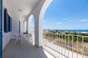 Een balkon met witte stoelen en een tafel, met uitzicht op een strand en stadsbeeld.