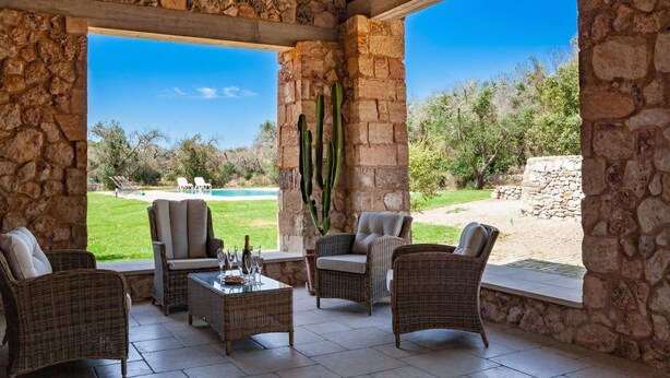 Une terrasse couverte, meublée de mobilier en osier, avec un mur en pierre et une vue sur une piscine et un jardin.