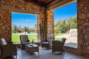Eine überdachte Terrasse mit Korbmöbeln, einer Steinmauer und Blick auf einen Pool und die Gartenlandschaft.