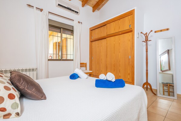 Una cama bien hecha con una colcha blanca, una toalla azul y una almohada marrón. Se observa un armario de madera y un espejo en la pared.