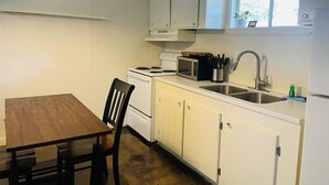 Eine kleine Küche mit weißen Schränken, einem Holztisch, einem schwarzen Stuhl, einer Mikrowelle, einem Herd und einem Spülbecken.