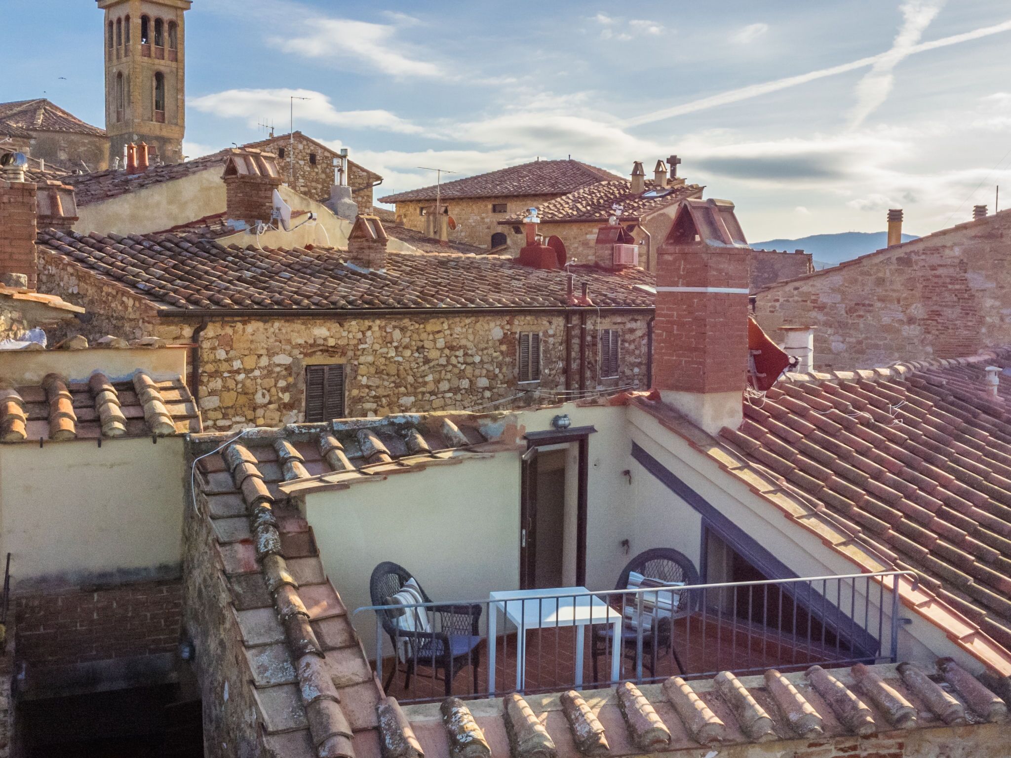 Una terrassa al terrat amb taula i cadires, edificis de pedra i una torre al fons.