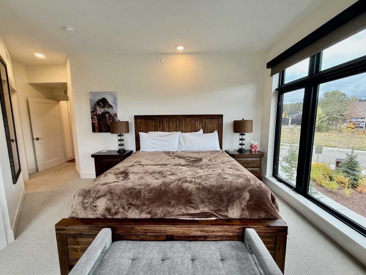 Un dormitorio con una cama grande, cabecera de madera, dos mesitas de noche con lámparas, un ventanal con vistas y una puerta que da a otra habitación.