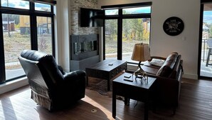 A modern living room with a stone fireplace, leather furniture, and large windows offering a view of the outdoors. - New Luxury 3-Bedroom Condo 1/2 Block to Main St (Winter Park)
