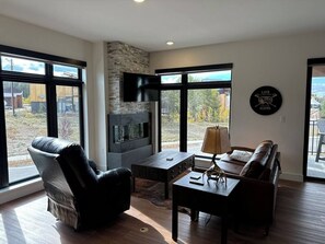A modern living room with a stone fireplace, leather furniture, and large windows offering a view of the outdoors. - New Luxury 3-Bedroom Condo 1/2 Block to Main St (Winter Park)