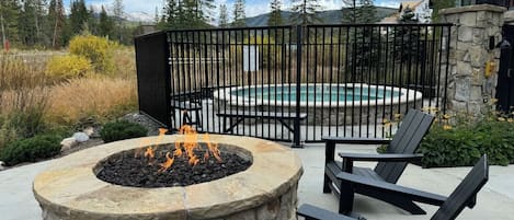 Un hogar de piedra con fuego, rodeado de sillas Adirondack negras, con una piscina y un edificio al fondo.