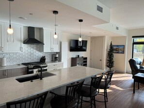 A modern kitchen with a central island, dining chairs, a coffee machine, and pendant lighting. - New Luxury 3-Bedroom Condo 1/2 Block to Main St (Winter Park)
