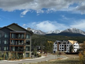 A multi-story building with balconies, a road in the foreground, and snow-capped mountains in the background.