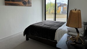 A bedroom with a large bed, a window with a view, and a lamp on a bedside table. - New Luxury 3-Bedroom Condo 1/2 Block to Main St (Winter Park)