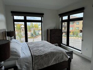 A bedroom with a bed, a dresser, and a window with a view of a parking lot and buildings.