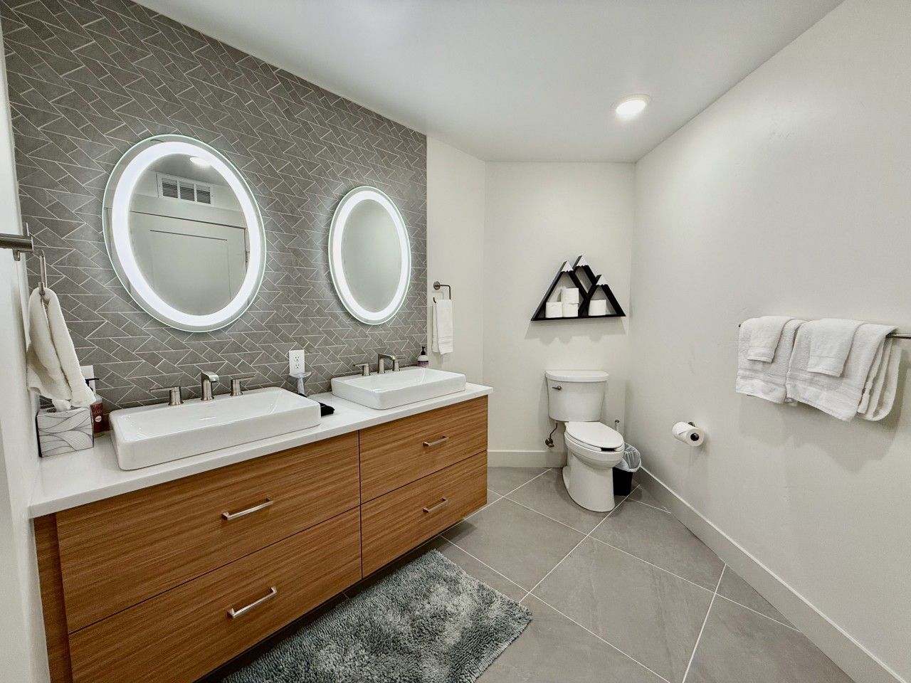 A modern bathroom with a double sink vanity, a toilet, and a mirror with built-in lighting.