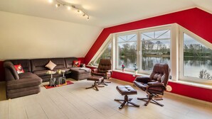 Un salon comprenant un canapé d’angle, deux fauteuils en cuir, une table basse en verre et une vue sur un lac, visible à travers de grandes fenêtres.