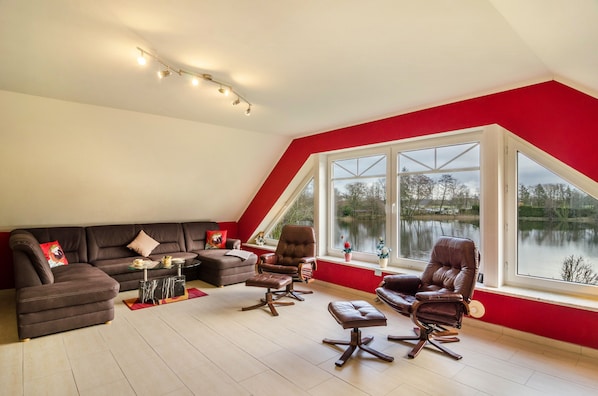 Un salon comprenant un canapé d’angle, deux fauteuils en cuir, une table basse en verre et une vue sur un lac, visible à travers de grandes fenêtres.