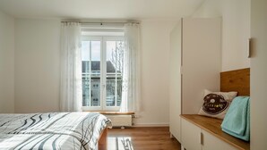 A bedroom with a bed, a window with curtains, a radiator, a wooden dresser, and a pillow with a speedometer design. - Apartment 'Windhager Seeblick' with Lake View, Balcony and Wi-Fi (Friedrichshafen)