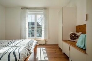 Une chambre à coucher avec un lit, une fenêtre avec des rideaux, un radiateur, une commode en bois et un oreiller orné d’un motif représentant un compteur de vitesse.