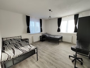 Una habitación con cama, escritorio y silla, con ventanas amplias y una lámpara de techo.