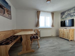 Una habitación con un banco de madera, una mesa de comedor, un radiador, un televisor y una ventana con cortinas.