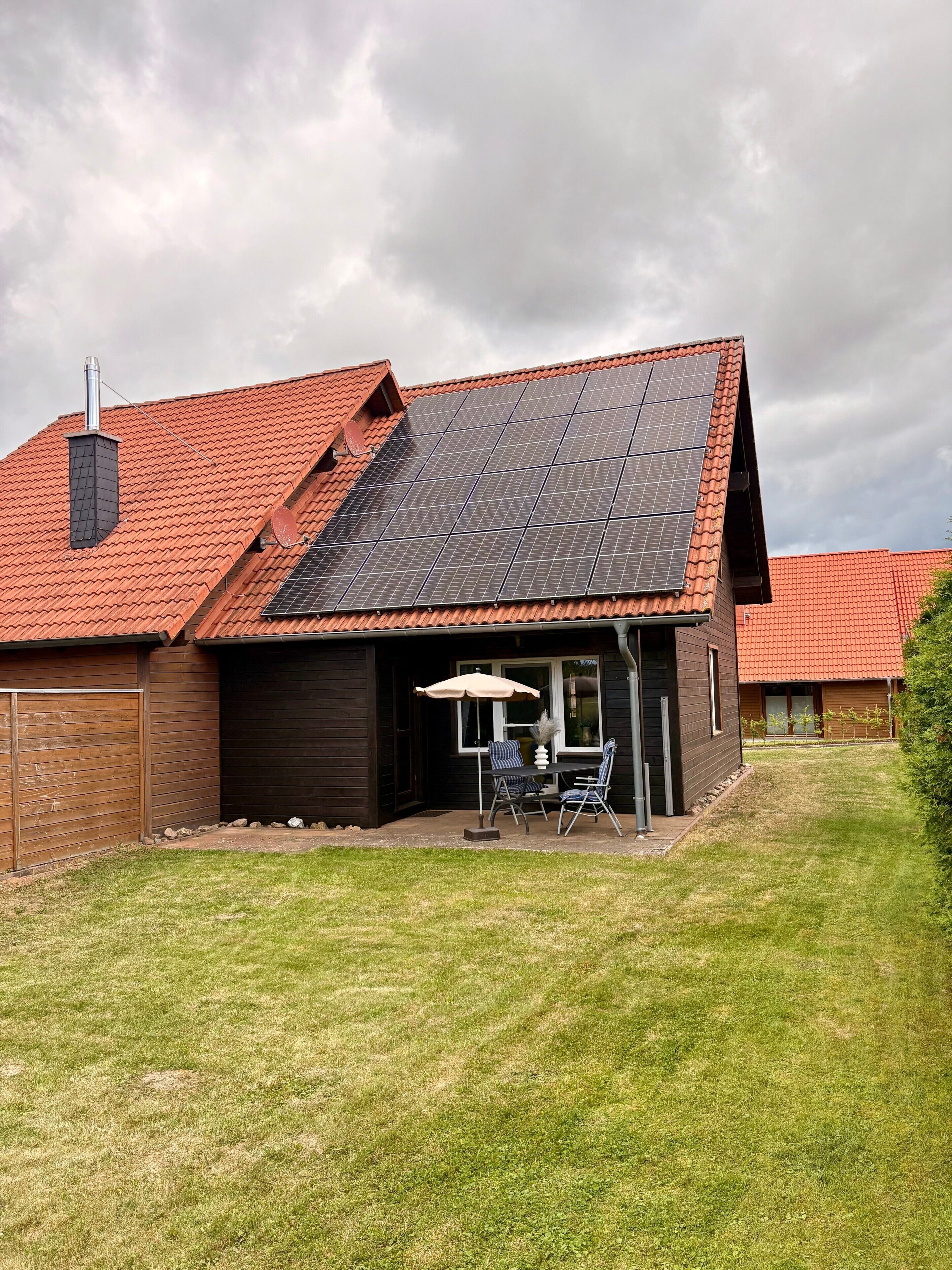 A house with a sloped roof, solar panels, a patio with chairs and an umbrella, and a fenced yard.