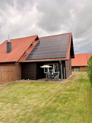 Ein Haus mit geneigtem Dach, Solaranlagen, einer Terrasse mit Stühlen und Sonnenschirm sowie einem eingezäunten Garten.