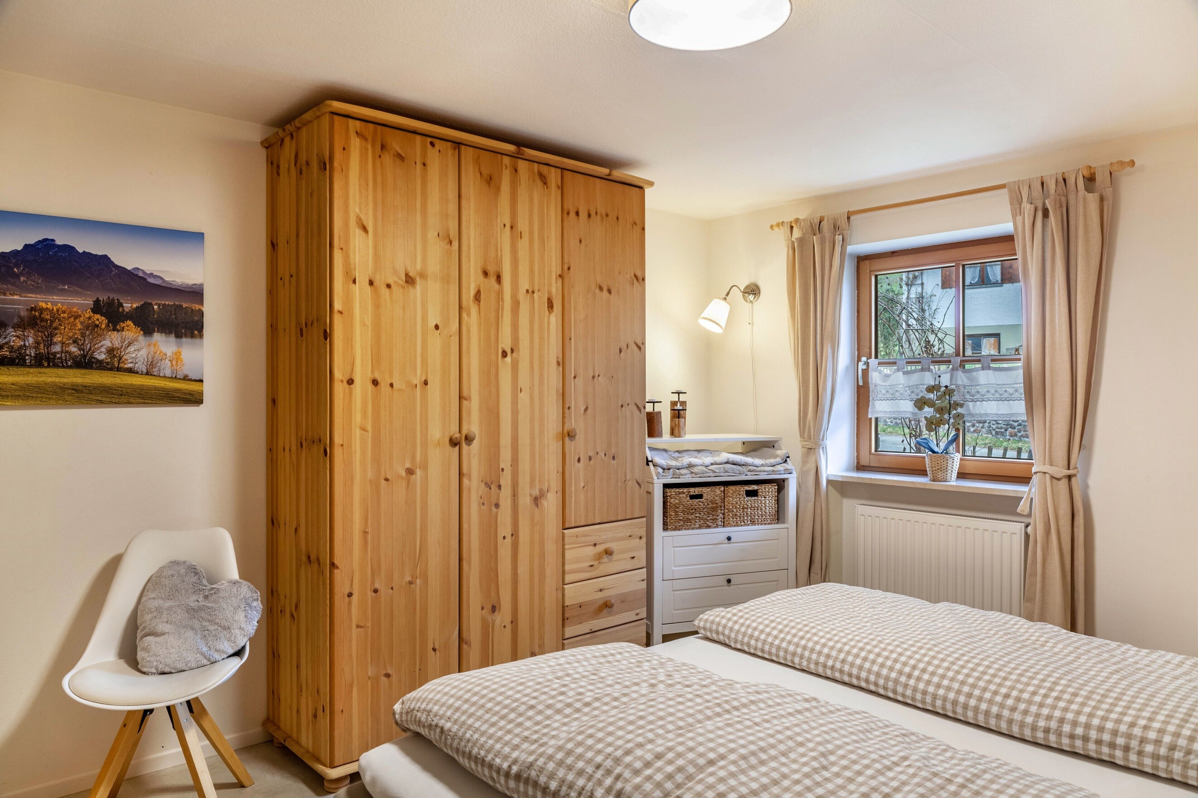 A bedroom with a wooden wardrobe, a bedside table with a lamp, a window with curtains, and a bed with checkered bedding.