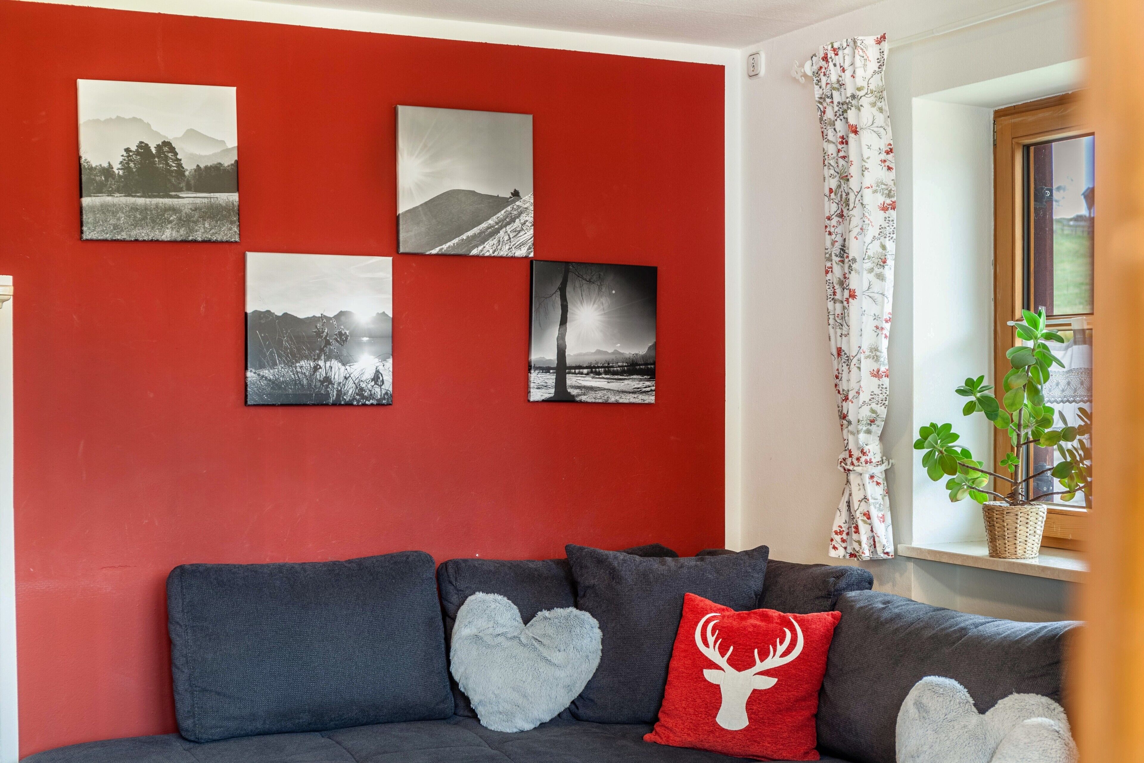 A living room with a red wall, a grey sofa with heart-shaped and deer-themed cushions, and a window with a plant.