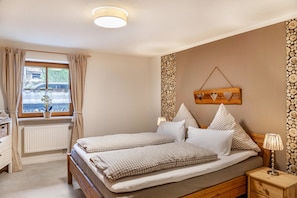 A bedroom with a double bed, a bedside table with a lamp, a window with curtains, a radiator, and a wall-mounted decoration. - Apartment "Mair - Bergblick" with Mountain View, Shared Terrace & Wi-Fi (Halblech)
