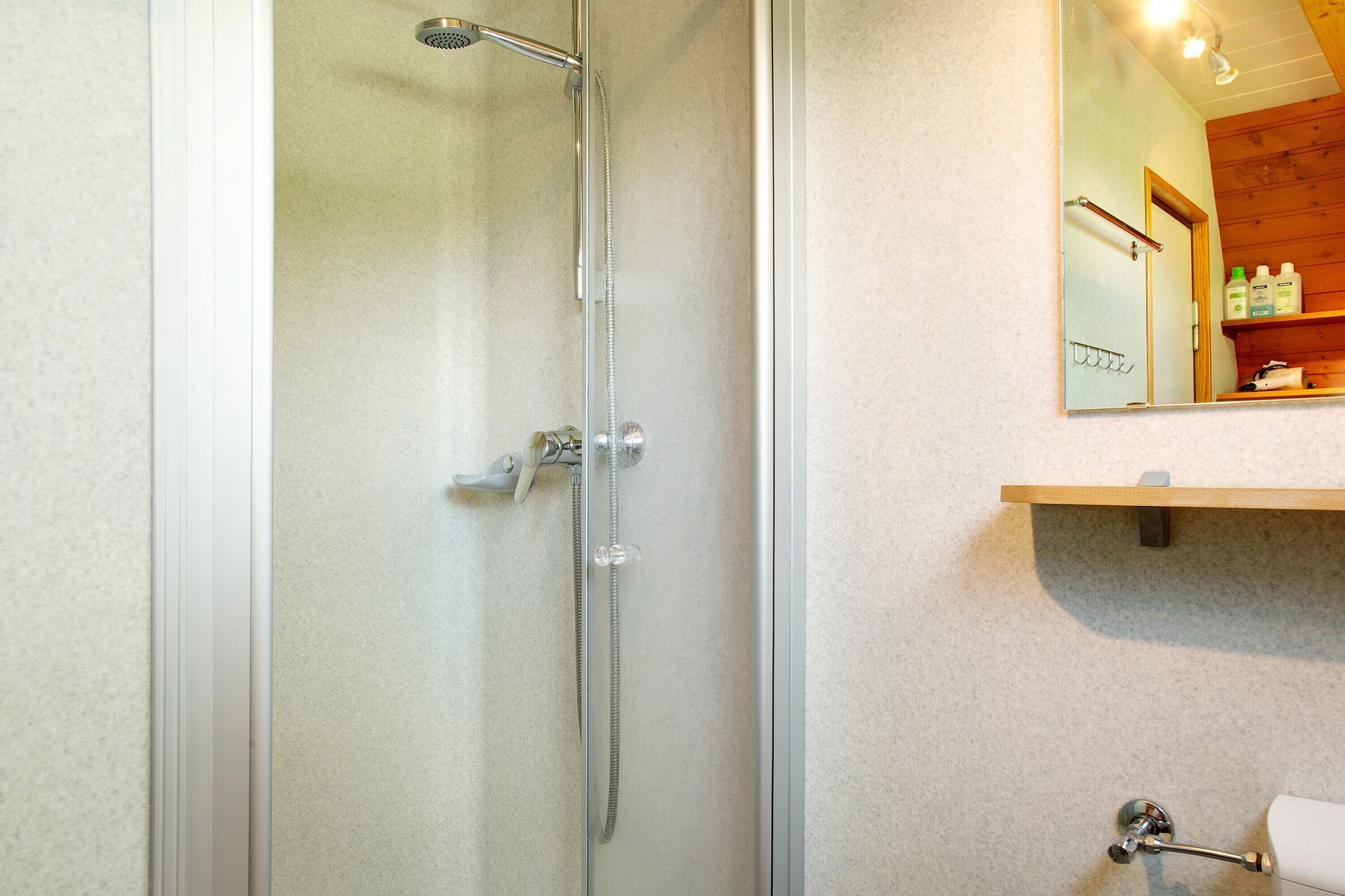 Une douche avec une porte en verre, une étagère en bois où sont rangés les articles de toilette et un miroir qui reflète la salle de bain.