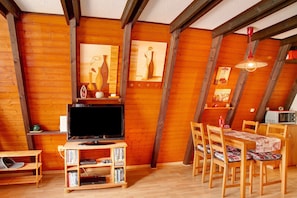 A wooden interior with a television, bookshelves, a dining table with chairs, and a ceiling fan.
