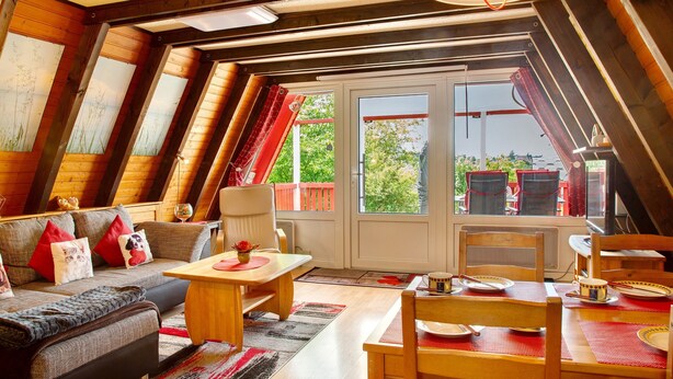 A cozy attic room with a sloped ceiling, a sofa with cushions, a dining table, chairs, and a view through a glass door. - Vacation Home "Villa Königsdorf Am See" with Lake View, Private Terrace & Wi-Fi (Waldkirchen)