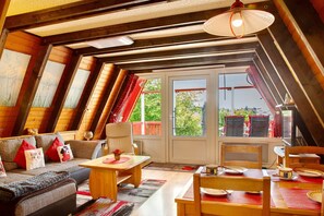 A cozy attic room with a sloped ceiling, a sofa with cushions, a dining table, chairs, and a view through a glass door. - Vacation Home "Villa Königsdorf Am See" with Lake View, Private Terrace & Wi-Fi (Waldkirchen)