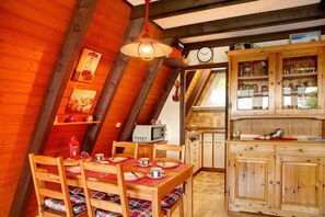 A wooden dining table with chairs, a kitchen area with wooden cabinets, a microwave, and a clock on the wall.