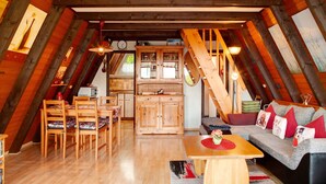 An attic room with wooden beams, a dining table with chairs, a sofa with cushions, and a kitchen area with cabinets and a microwave. - Vacation Home "Villa Königsdorf Am See" with Lake View, Private Terrace & Wi-Fi (Waldkirchen)