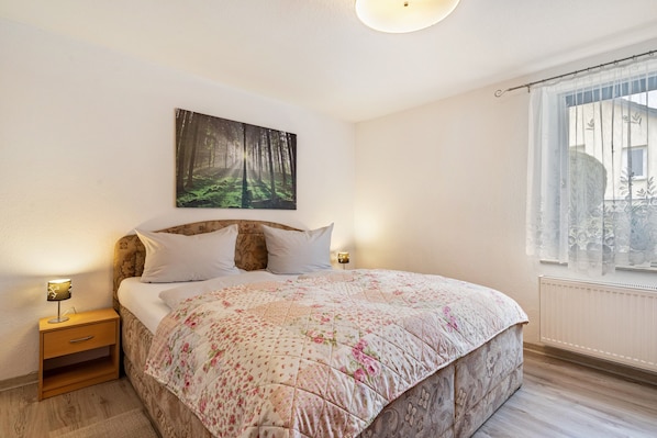 A bedroom with a bed, a nightstand, a wall-mounted painting, and a window with a curtain.