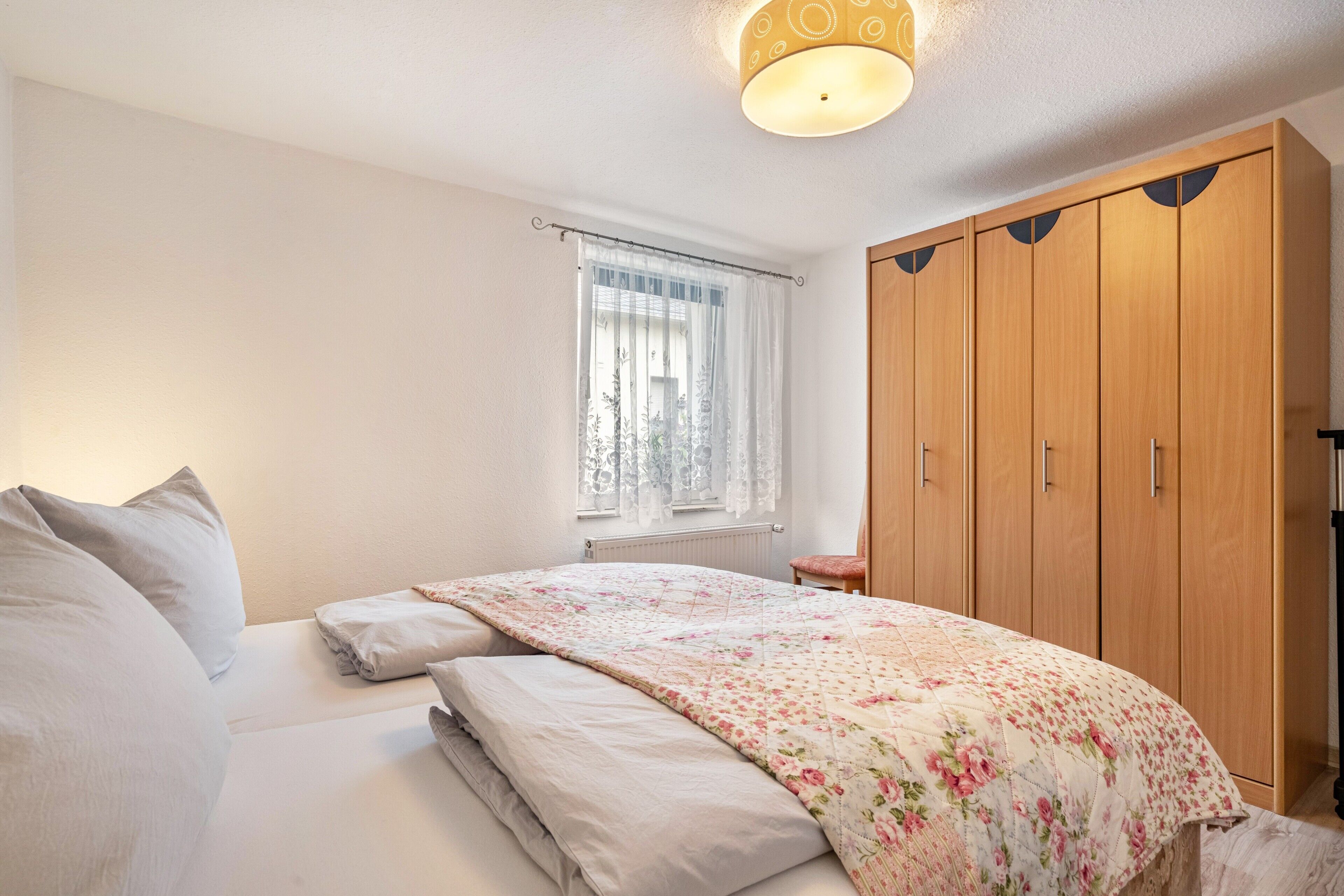 Ein Schlafzimmer mit einem Bett, einem Fenster mit Vorhängen, einem Holzschrank und einer Deckenleuchte.