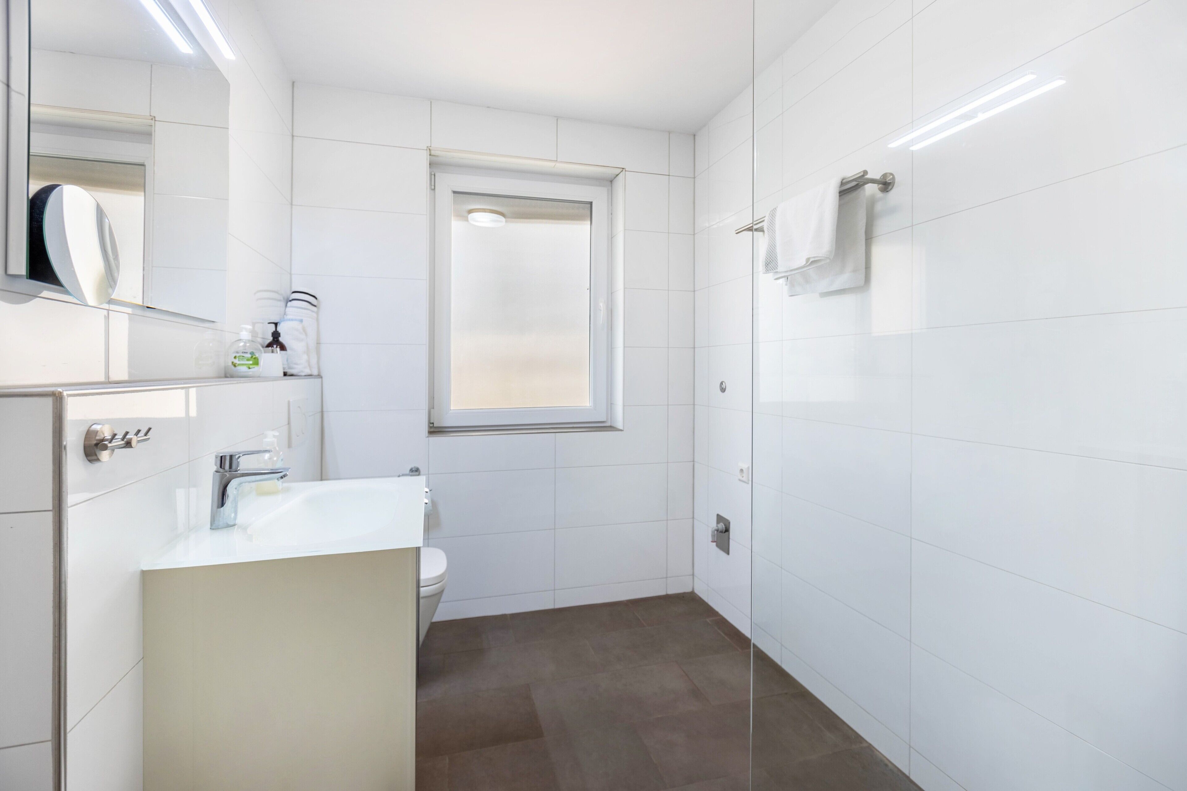 A modern bathroom with a white sink, mirror, and toilet, a glass shower enclosure, and a window.