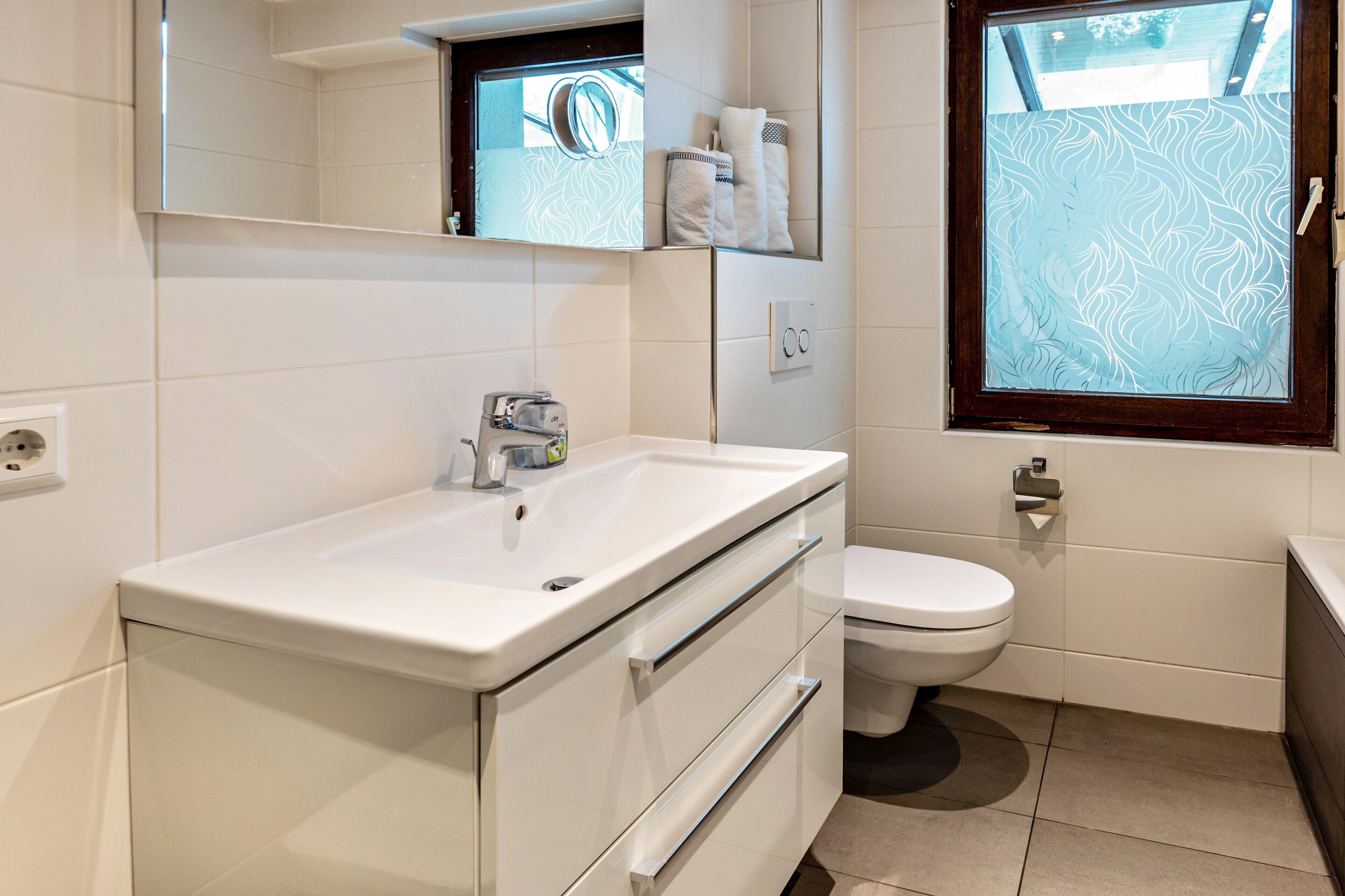 A modern bathroom with a white sink, a toilet, and a window with frosted glass.